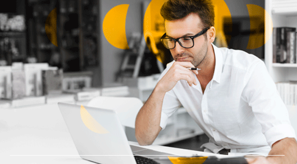 Un hombre con gafas mira la pantalla de una computadora portátil, sosteniendo un bolígrafo, en una oficina moderna con una estantería.