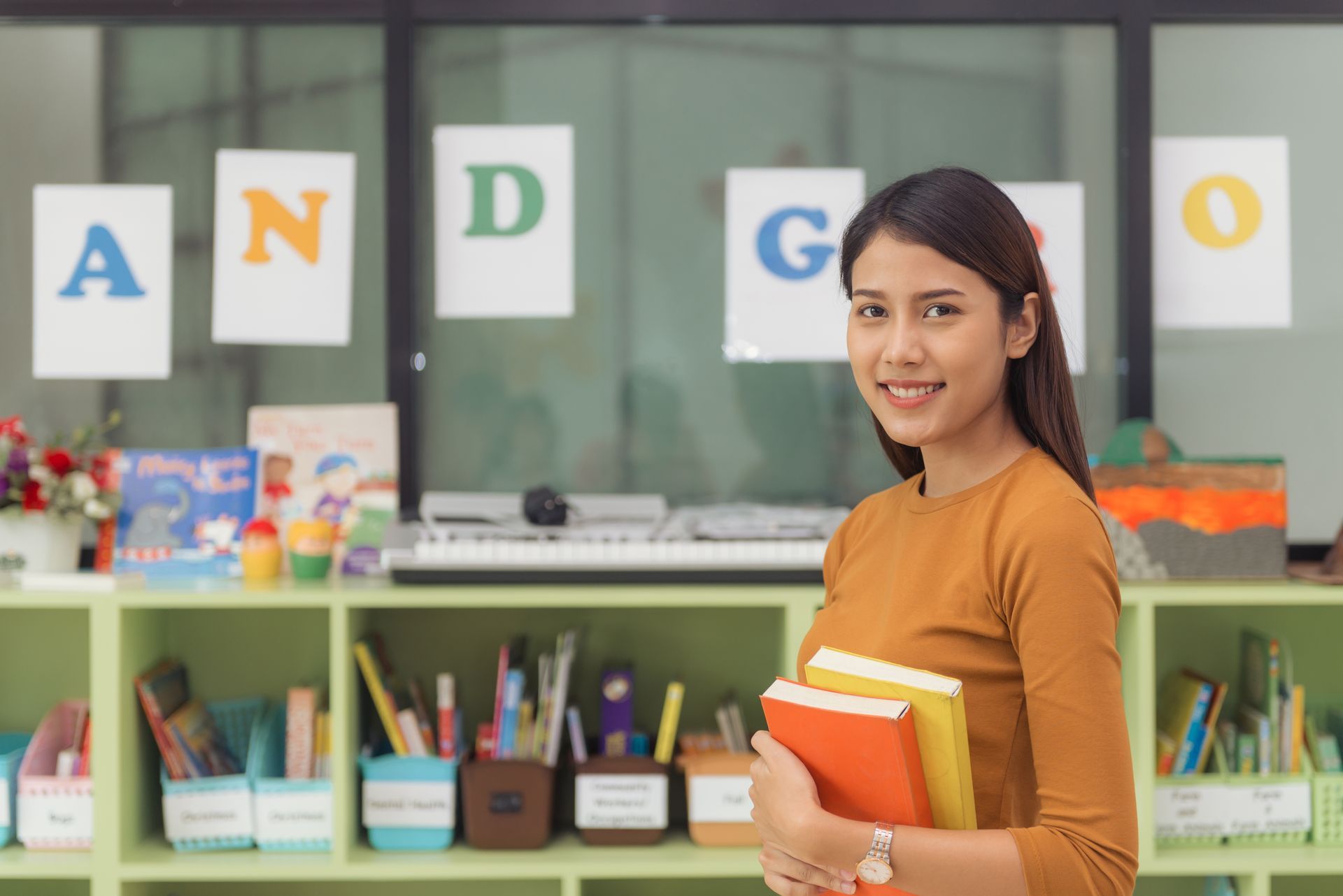 Profesora sonriendo, sosteniendo libros en un aula colorida con carteles de
