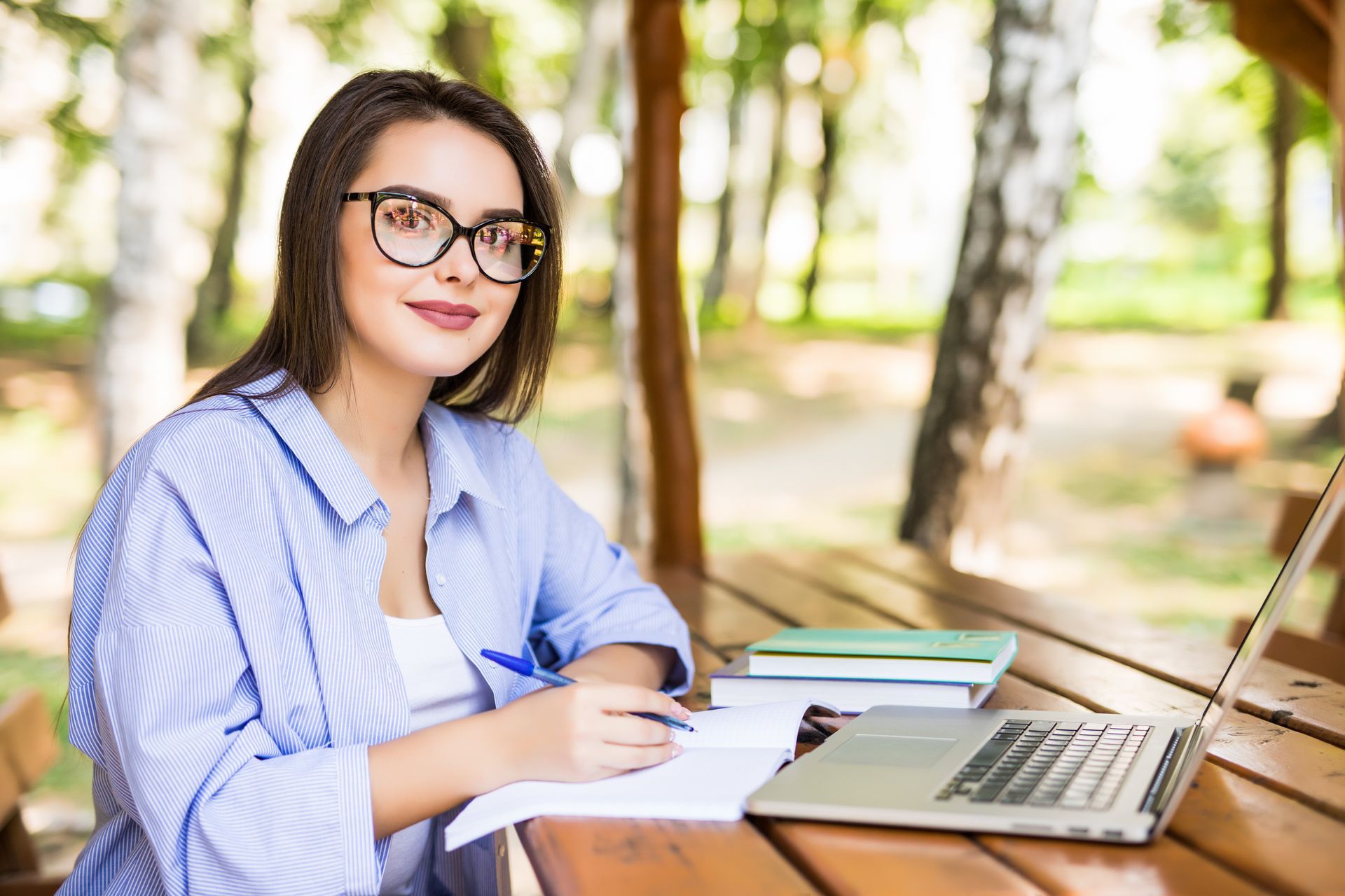 Mujer con gafas en una mesa al aire libre, escribiendo con una computadora portátil y libros cerca.
