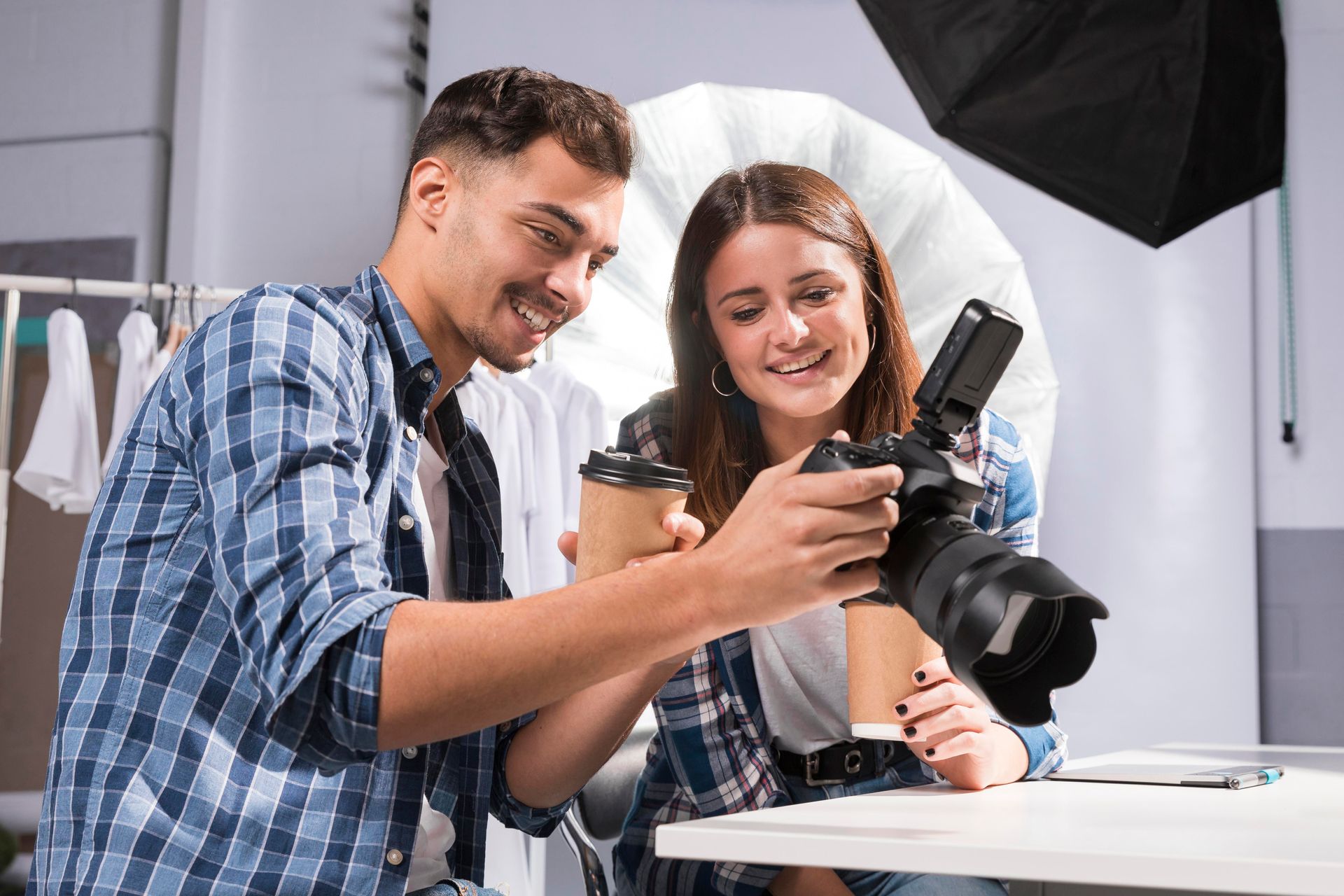 Dos personas sonriendo mirando a la cámara. Un hombre sostiene un café. Estudio con perchero y equipo de iluminación.