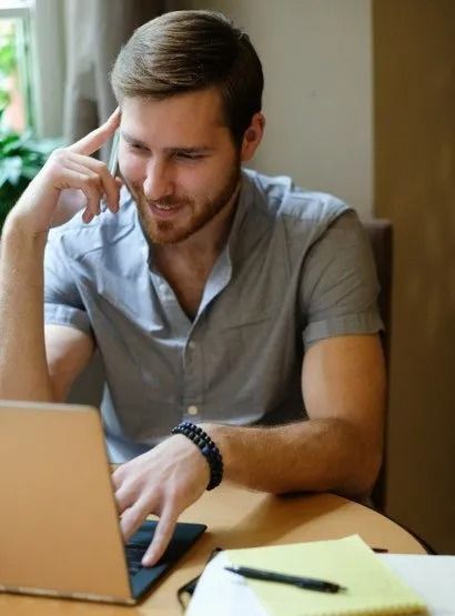 Hombre con barba, mirando una computadora portátil, sonriendo, apoyando su mano sobre su cabeza.