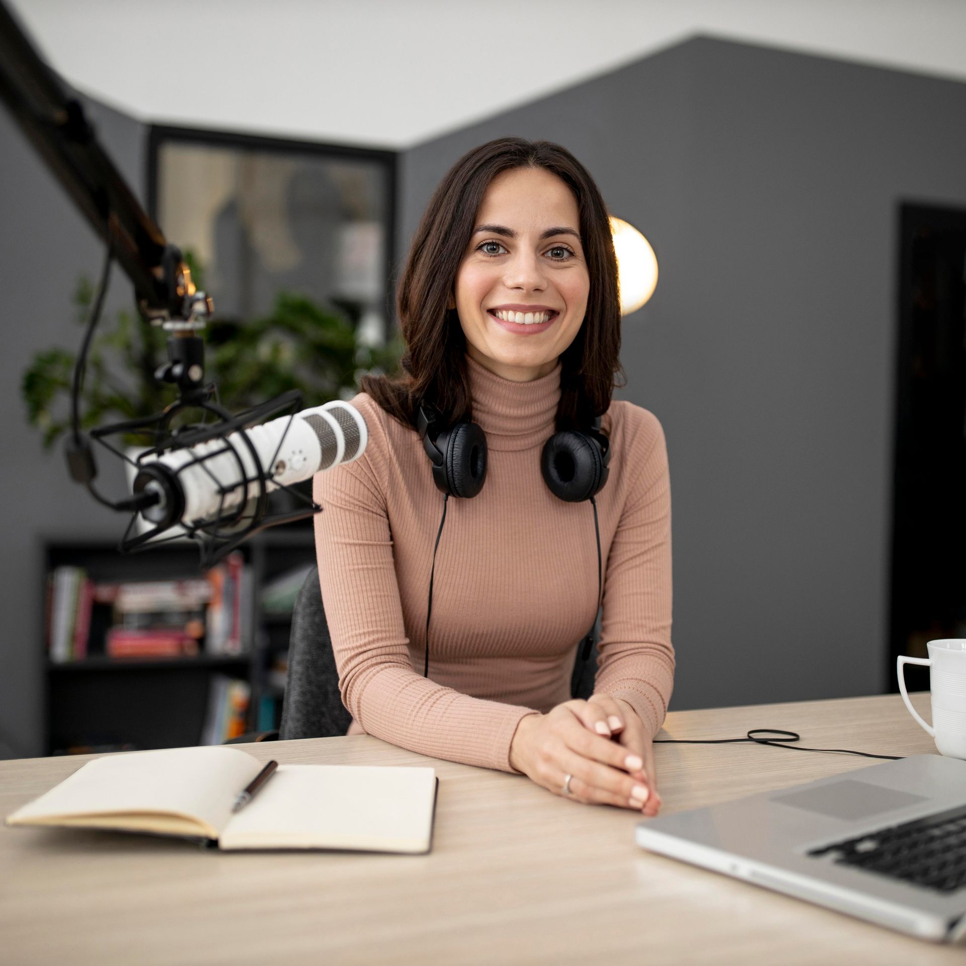 Mujer sonriendo en un escritorio con micrófono, auriculares y computadora portátil, en un entorno de estudio en casa.