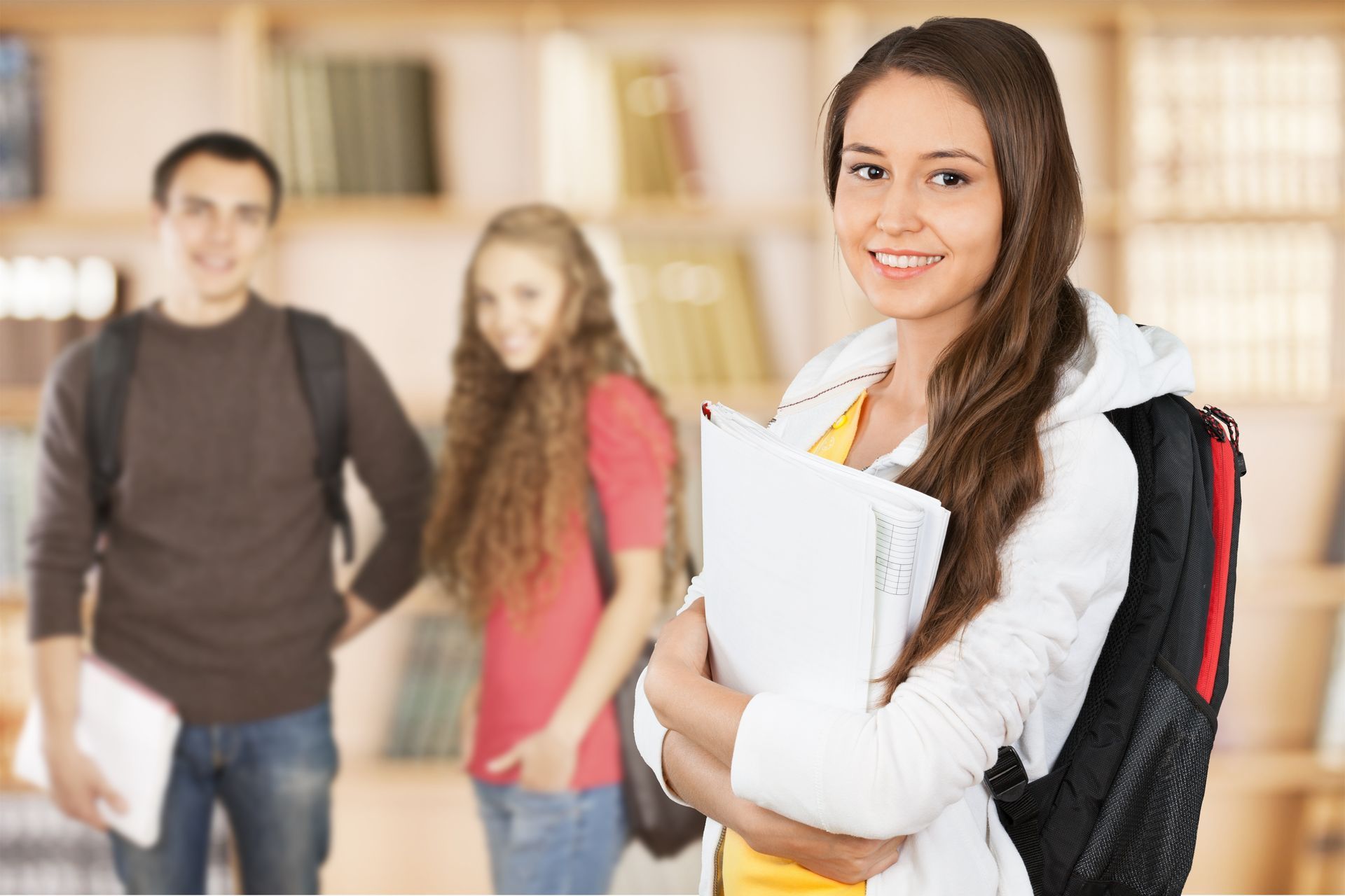 Estudiante sonriendo a la cámara, sosteniendo libros, con dos compañeros de clase borrosos en una biblioteca.