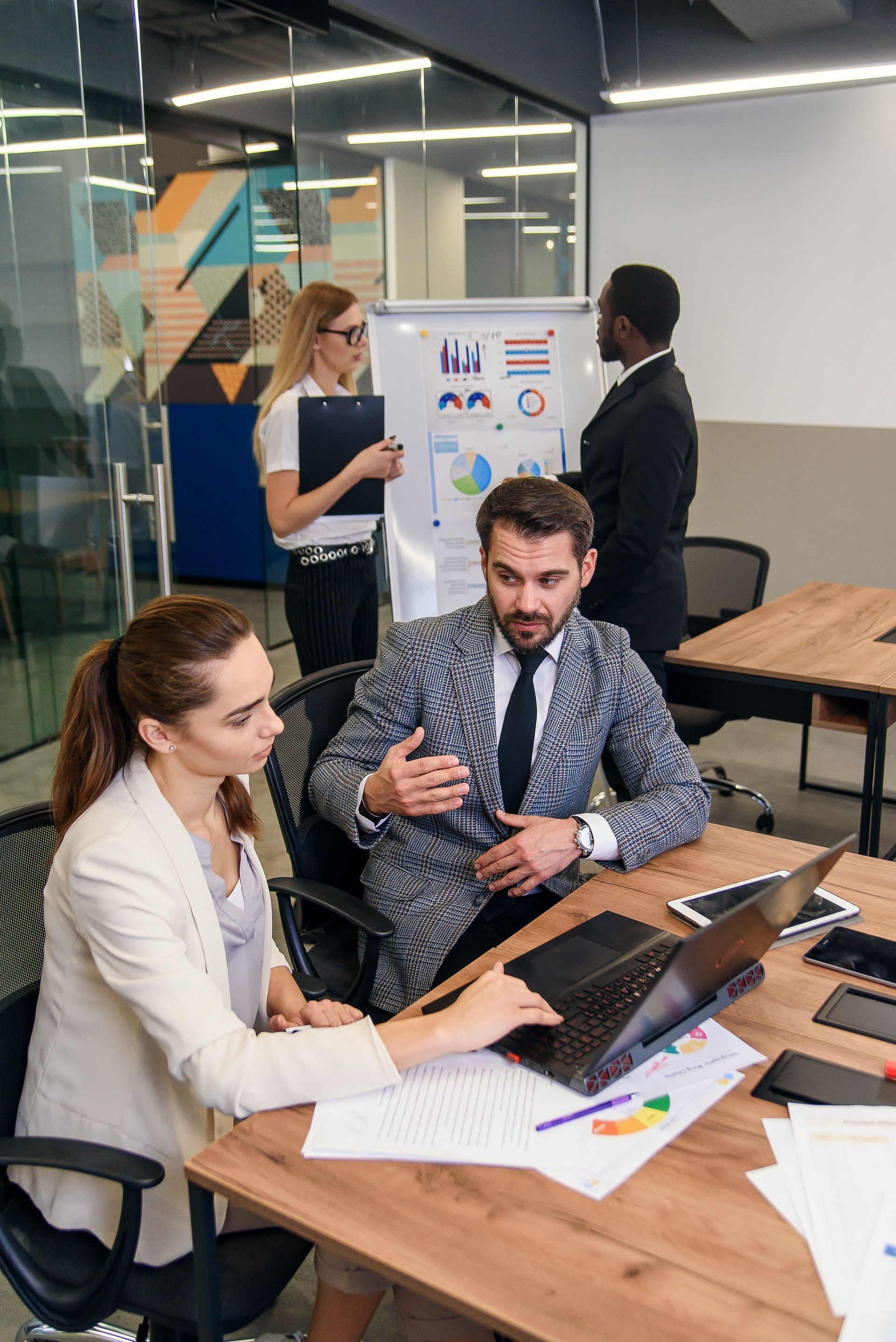 Reunión de negocios: colegas alrededor de una mesa con una computadora portátil y documentos; dos personas en una pizarra en el fondo.