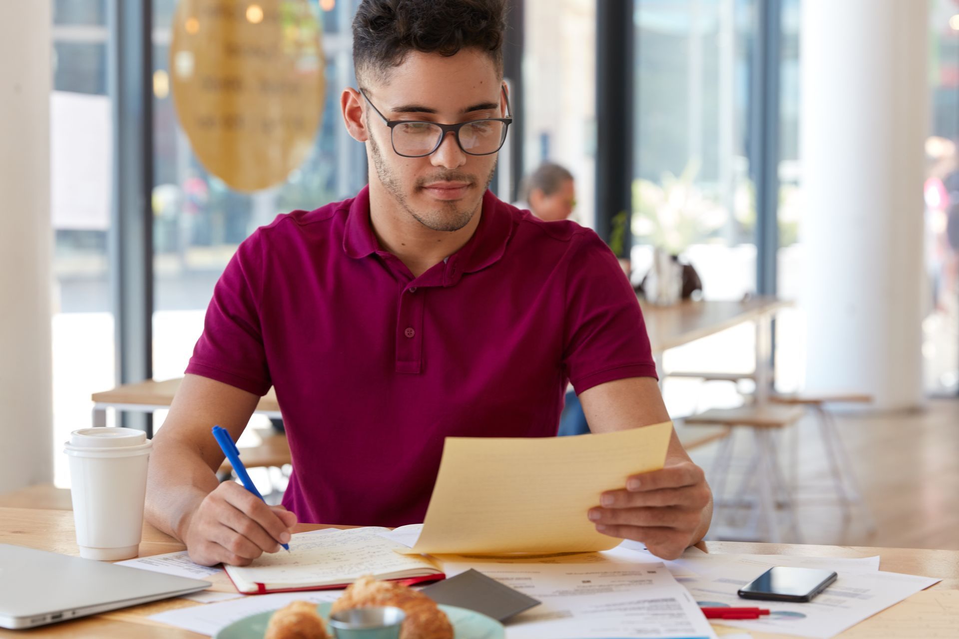 Hombre con gafas, camisa morada, escribiendo y mirando documentos en una mesa de un café.