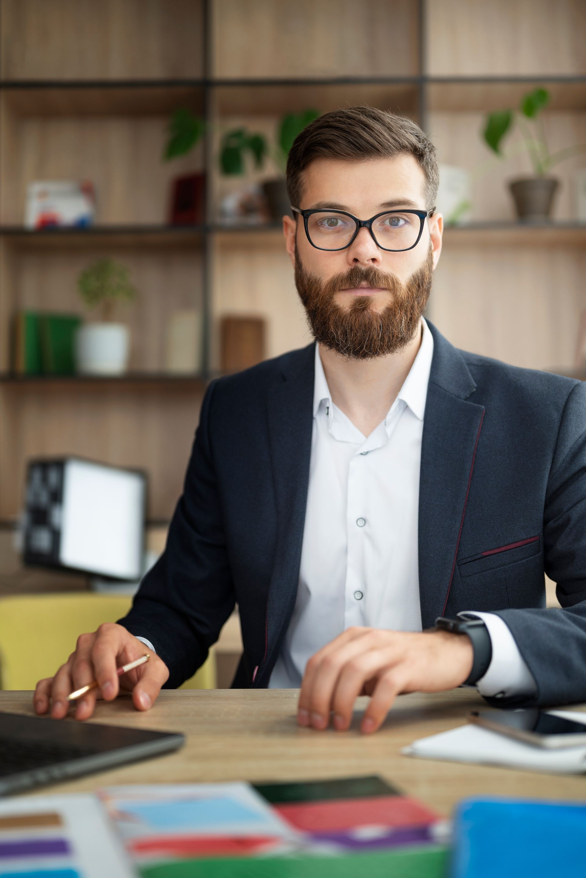 Hombre de traje, gafas y barba sentado en un escritorio, mirando a la cámara. Oficina con estanterías.