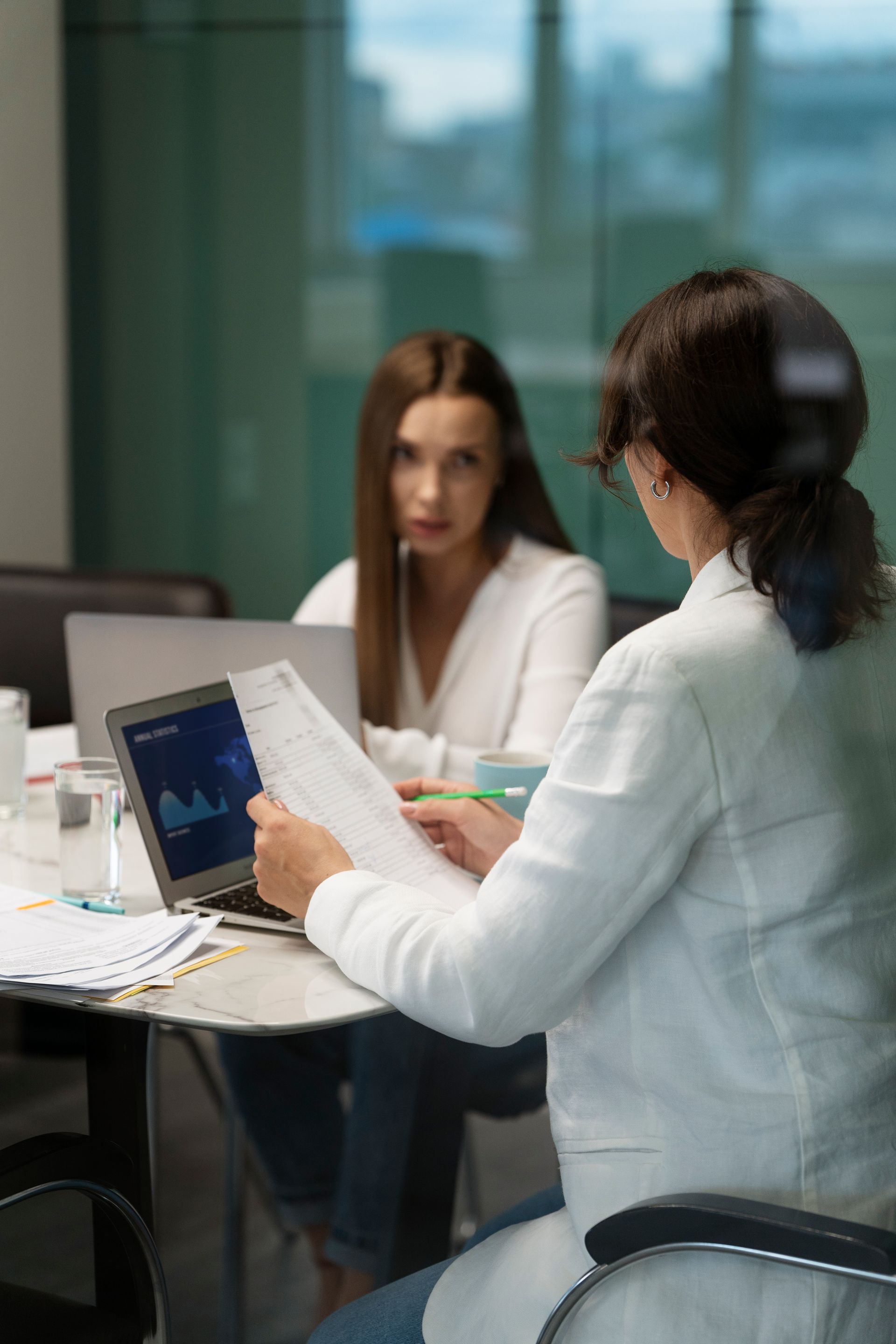 Dos mujeres en una reunión, una mirando papeles, la otra frente a una computadora portátil con gráficos, en un entorno de oficina moderno.