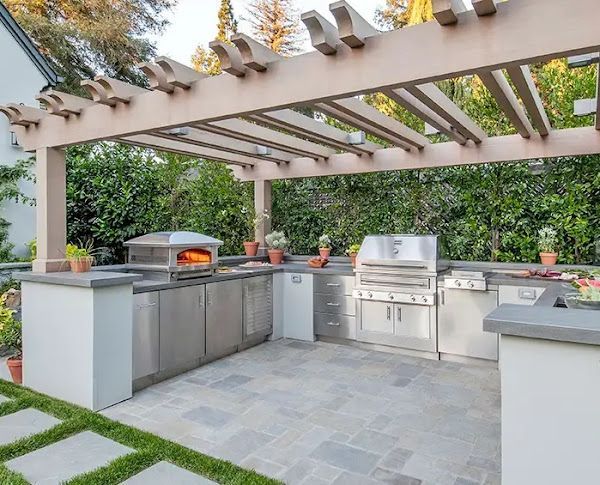 An outdoor kitchen with stainless steel appliances and a pizza oven under a pergola.