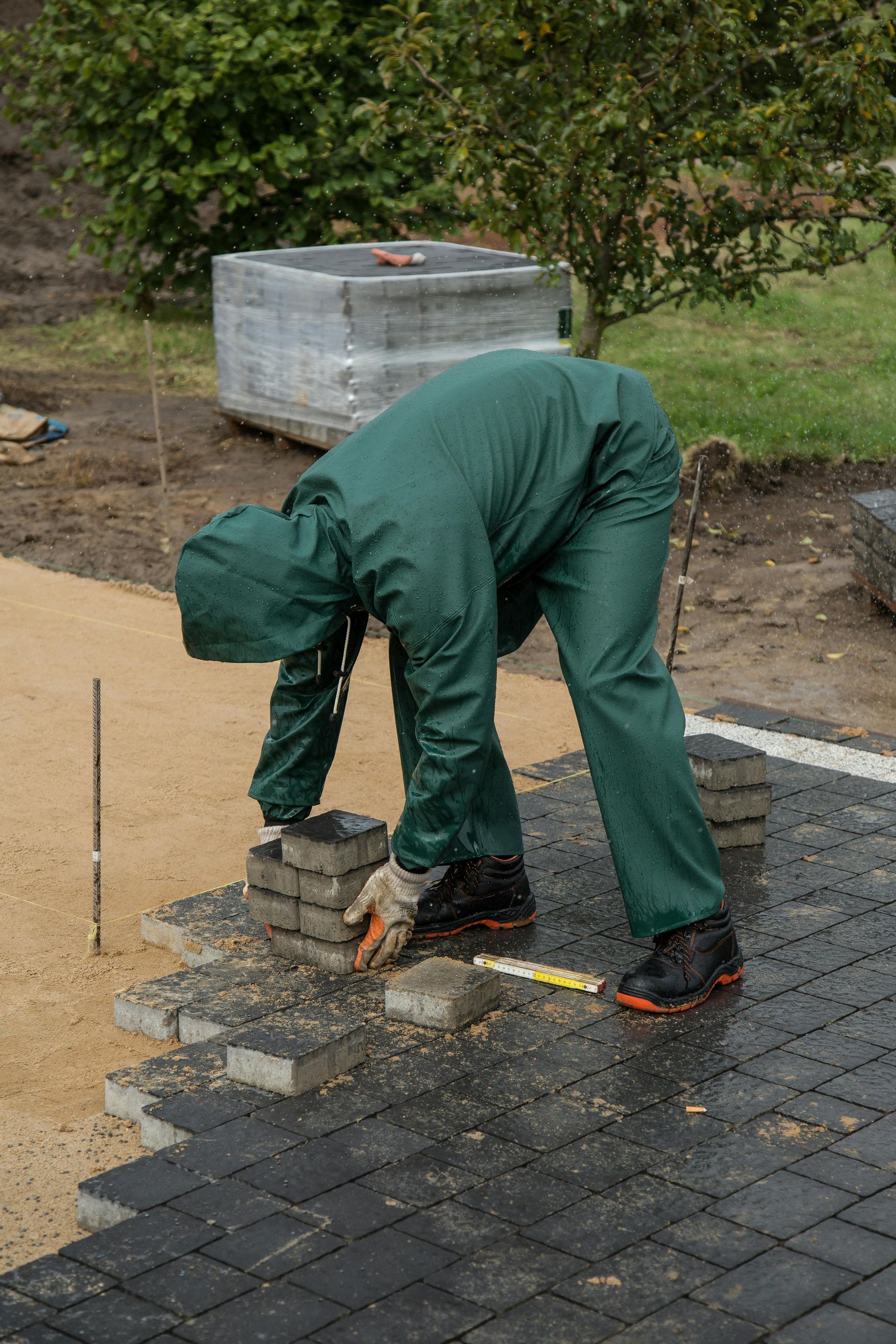A man in a green suit is laying bricks on a driveway.