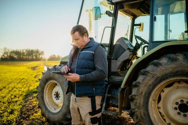 A farmer is standing besides a tractor and using a digital tablet in the field.