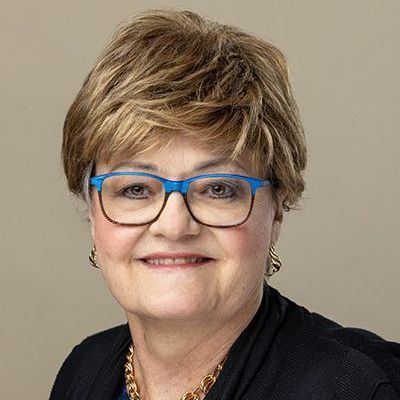 A woman wearing glasses and a blue shirt is smiling in front of a brick wall.