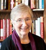 A woman wearing glasses and a purple scarf is smiling in front of a bookshelf.