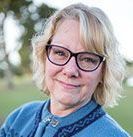 A woman wearing glasses and a blue sweater is smiling for the camera.