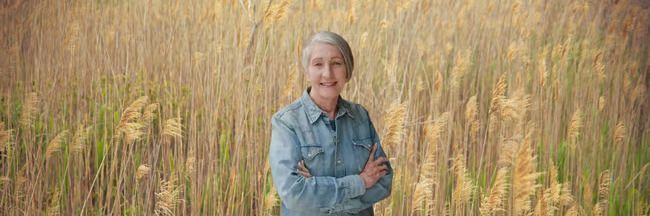 An elderly woman is standing in a field with her arms crossed.