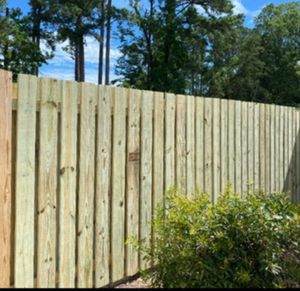 A newly installed light-wood privacy fence standing in front of green trees under a bright blue sky.