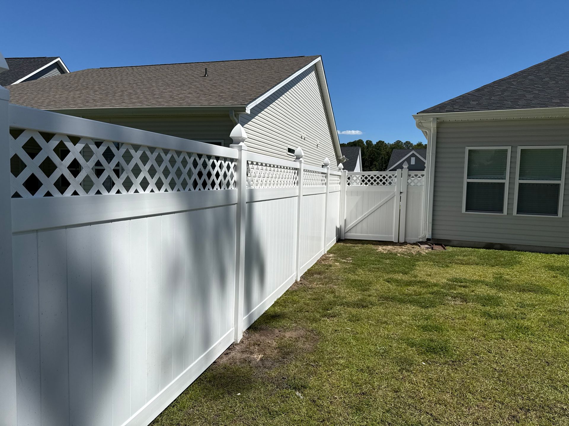 A white vinyl fence with a lattice top border runs alongside a light-colored house in a grassy yard under a blue sky.