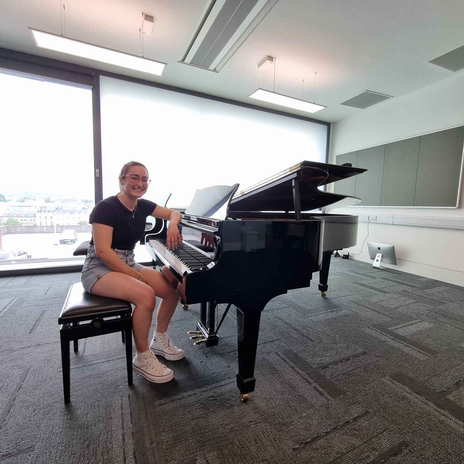 Person seated at a grand piano in a bright room with large windows, smiling at the camera.