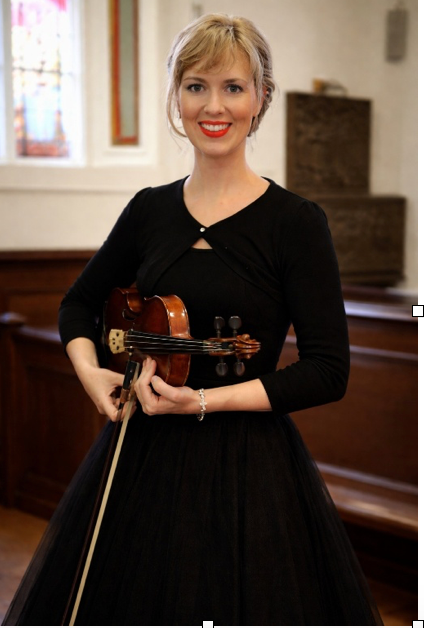 Woman in a black dress holding a violin indoors, smiling at the camera.