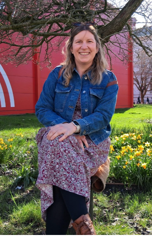 Woman sitting outdoors in a denim jacket and floral dress, smiling in a sunny garden.