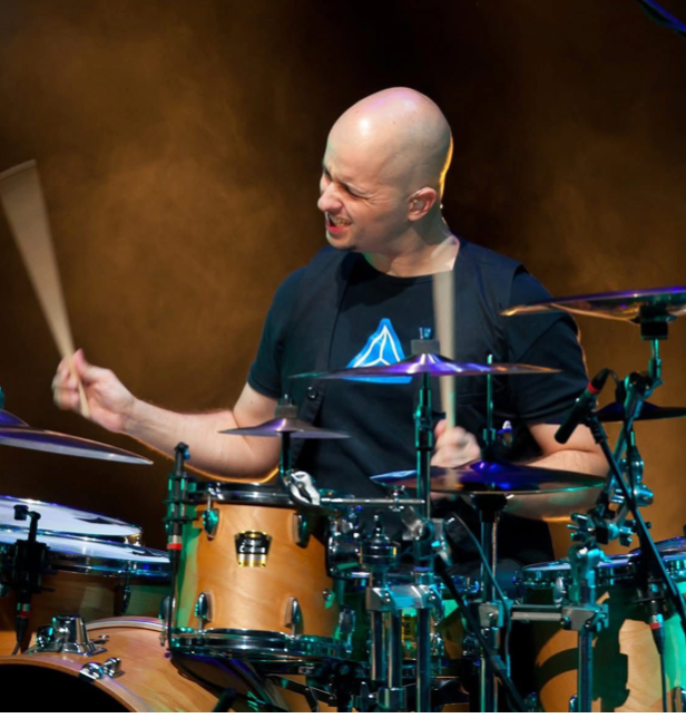 Drummer playing onstage, smiling under warm lights, with a blue graphic T-shirt and drum kit visible