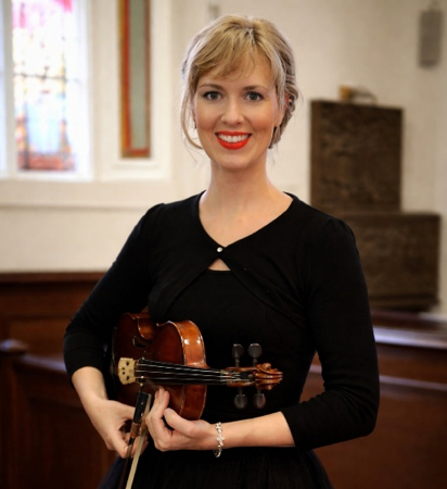 Woman holding a violin in a church interior, wearing a black top and smiling.