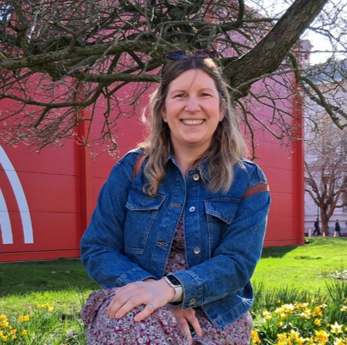 Smiling woman in a denim jacket sitting outdoors in front of a red building and flowering tree.