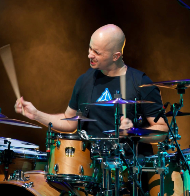 Drummer playing an amber drum kit on stage, smiling under warm concert lights
