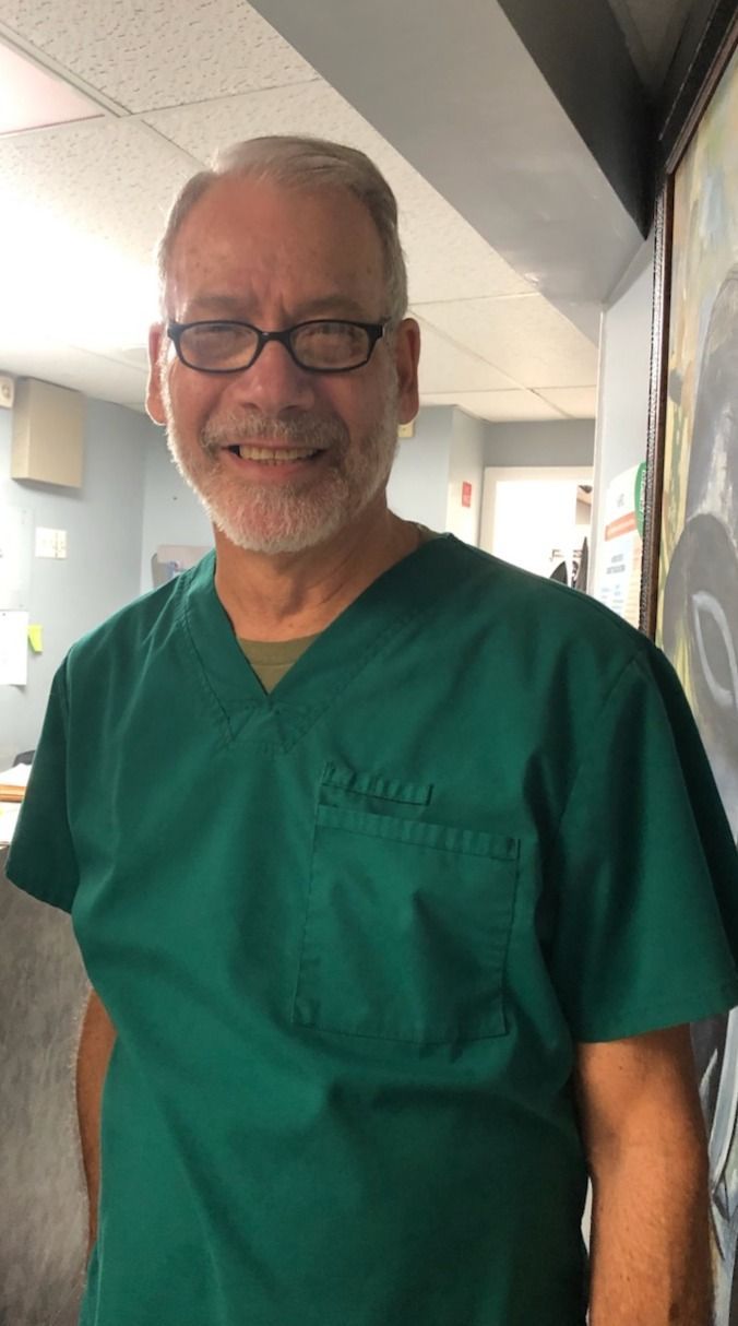 Man in green scrubs, glasses, smiles at camera; indoors, bright.