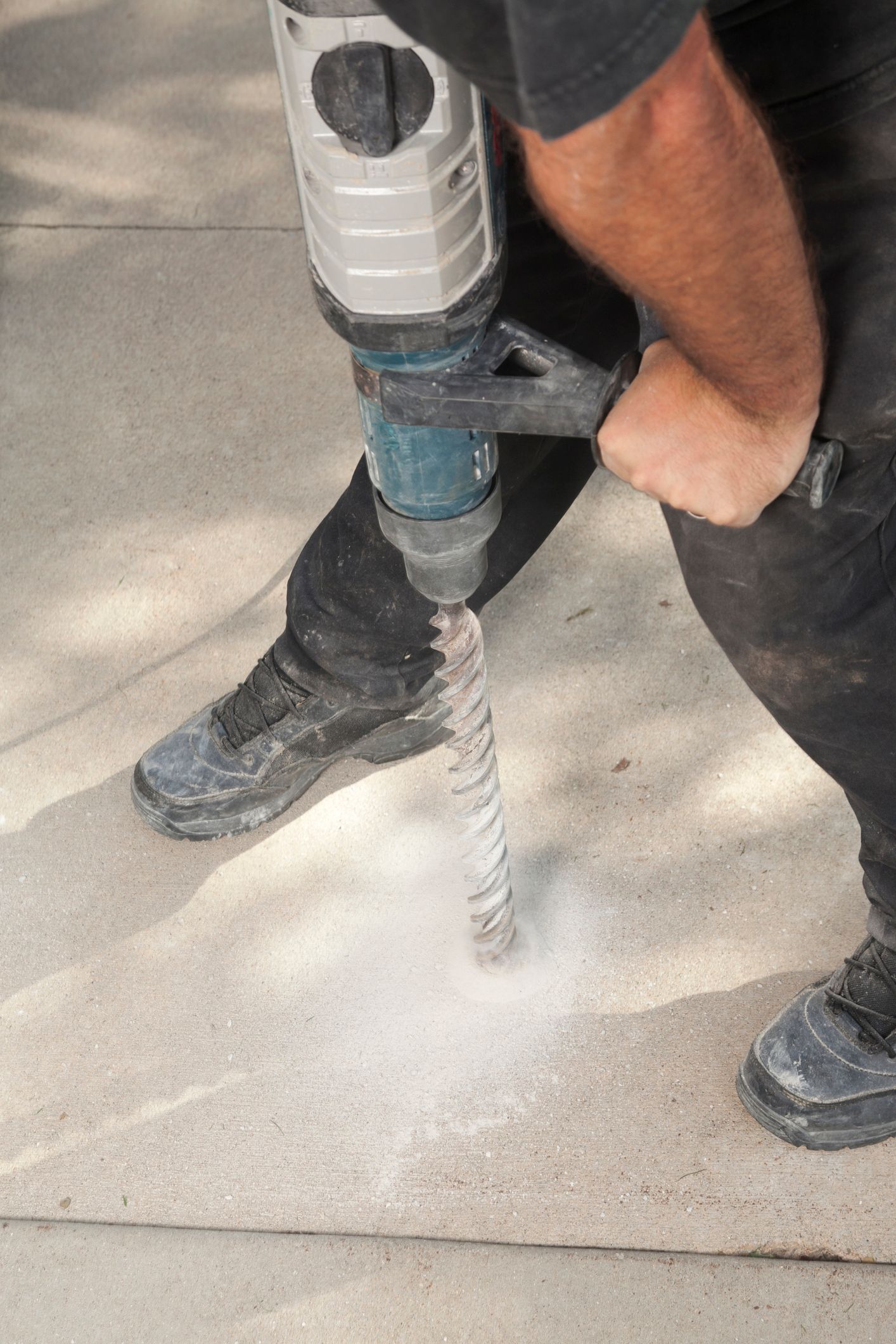 A worker is drilling a hole in the concrete driveway with a hammer drill tool for mudjacking. A worker is drilling a hole in the concrete driveway with a hammer drill tool for mudjacking.