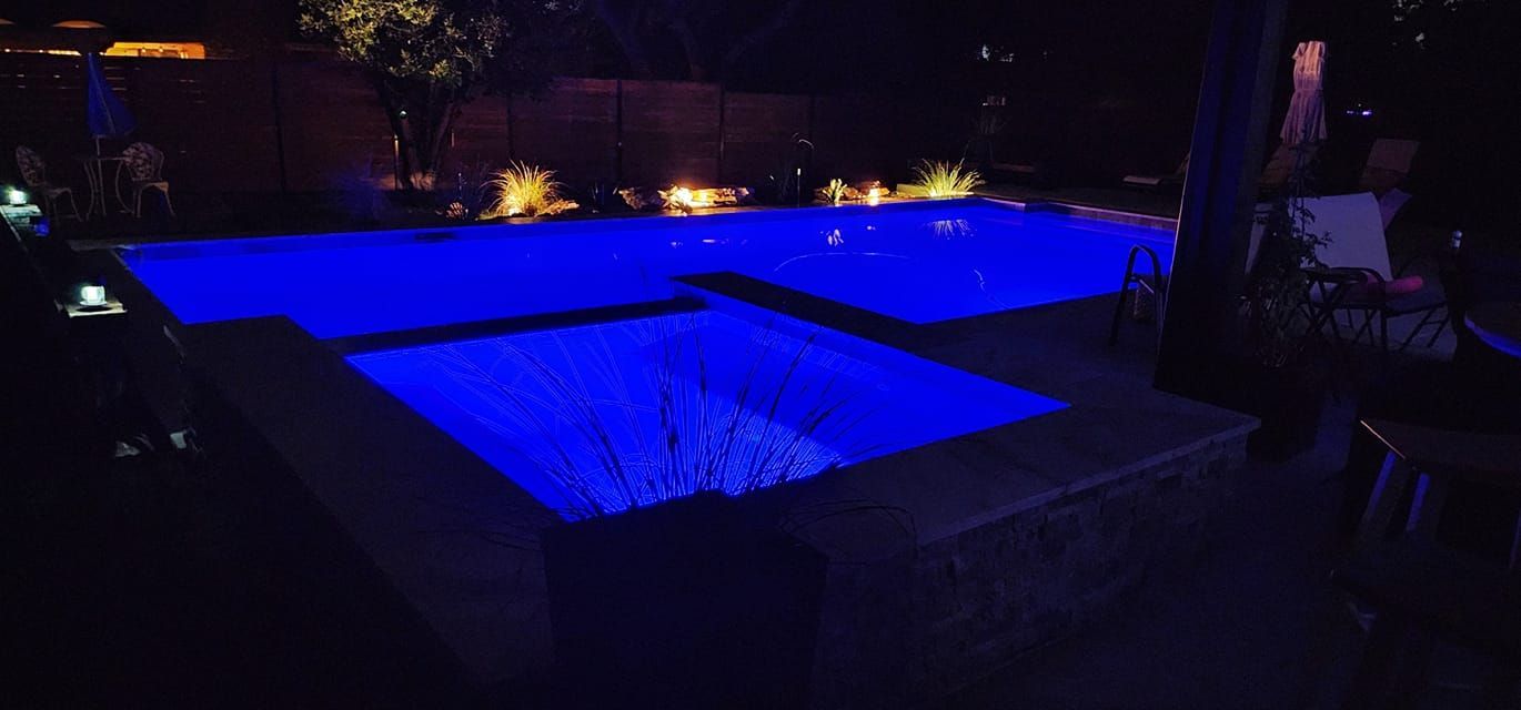 A glowing blue swimming pool and attached hot tub at night, surrounded by subtle outdoor landscape lighting.