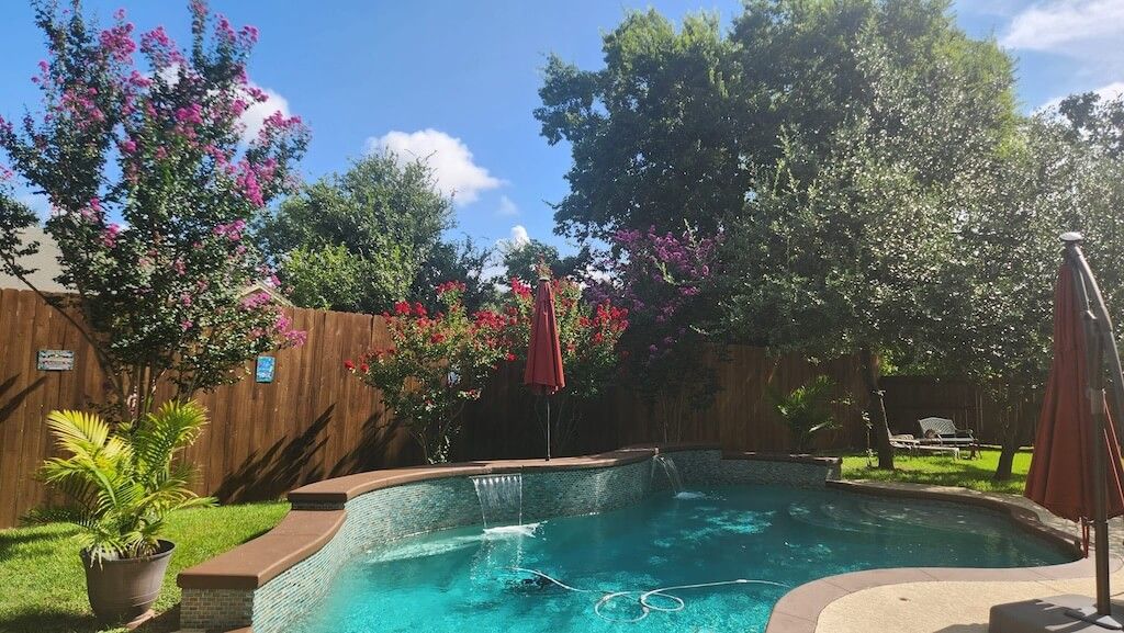 A sunny backyard features a turquoise swimming pool with a stone waterfall, surrounded by green trees and a wooden fence.