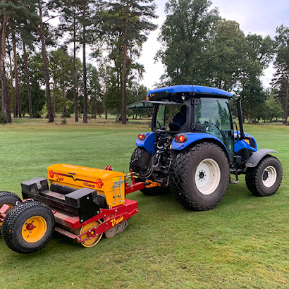 A blue tractor is pulling a yellow and black machine on a golf course.