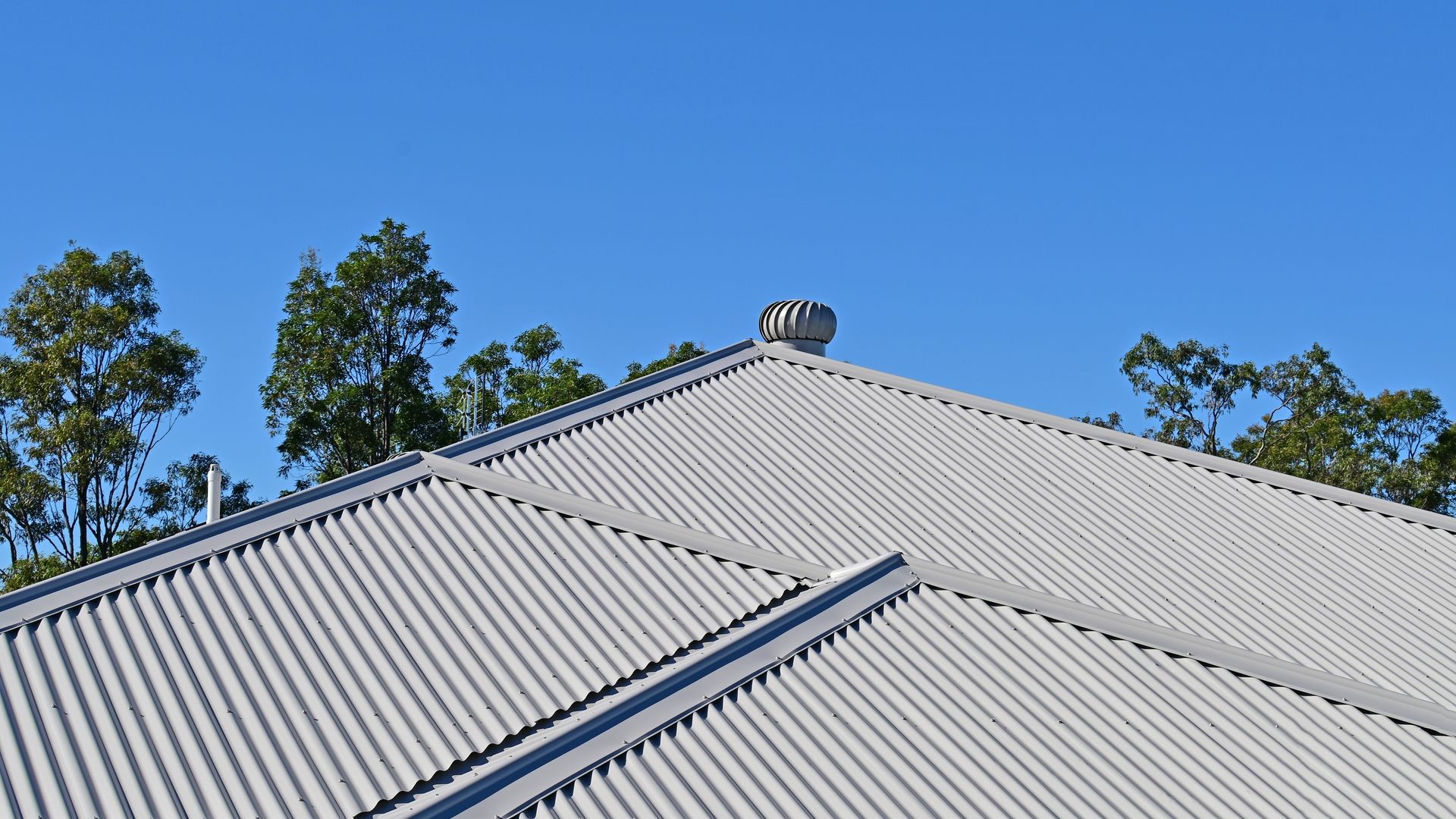 Gray corrugated metal roof with vent against a clear blue sky, trees in the background — C&A Roofing In Tweed Heads, NSW
