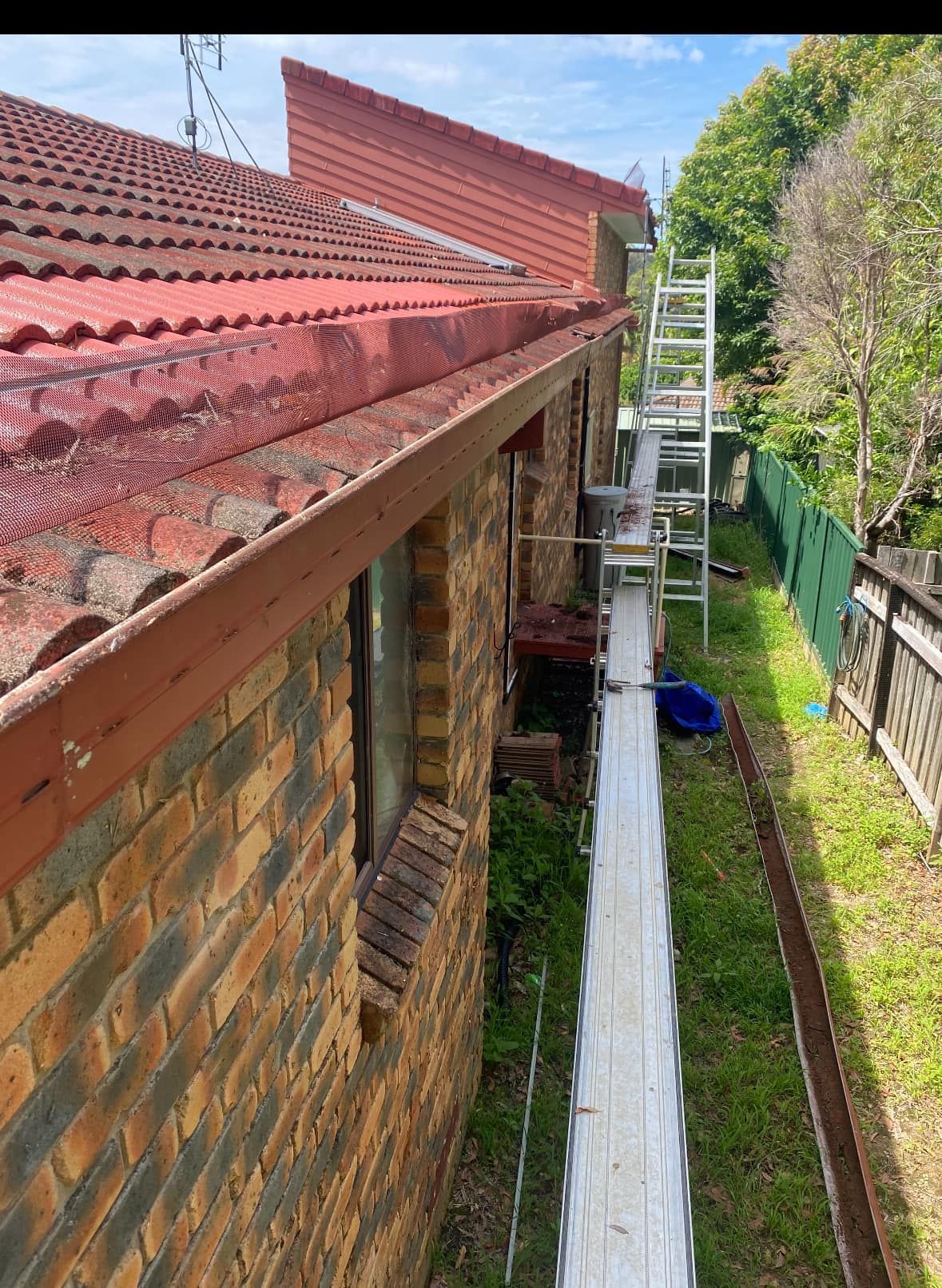 Side view of a brick house with a red tile roof and a ladder set up for gutter work.