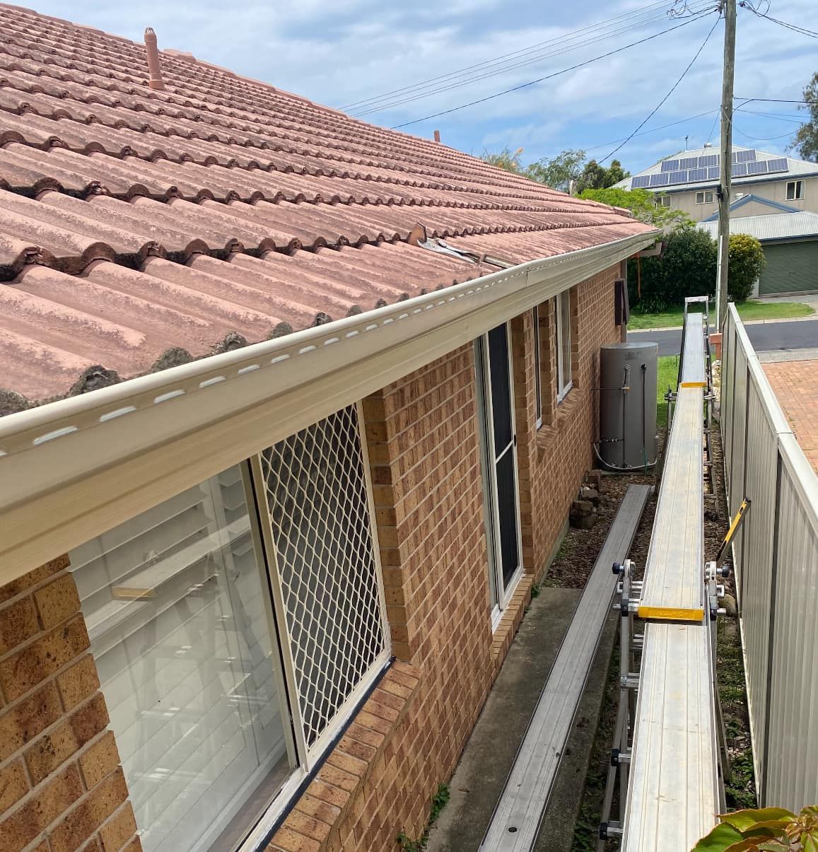 Red-tiled roof of a brick house, light-colored gutters, and a water heater next to windows — C&A Roofing in Boambee East, NSW