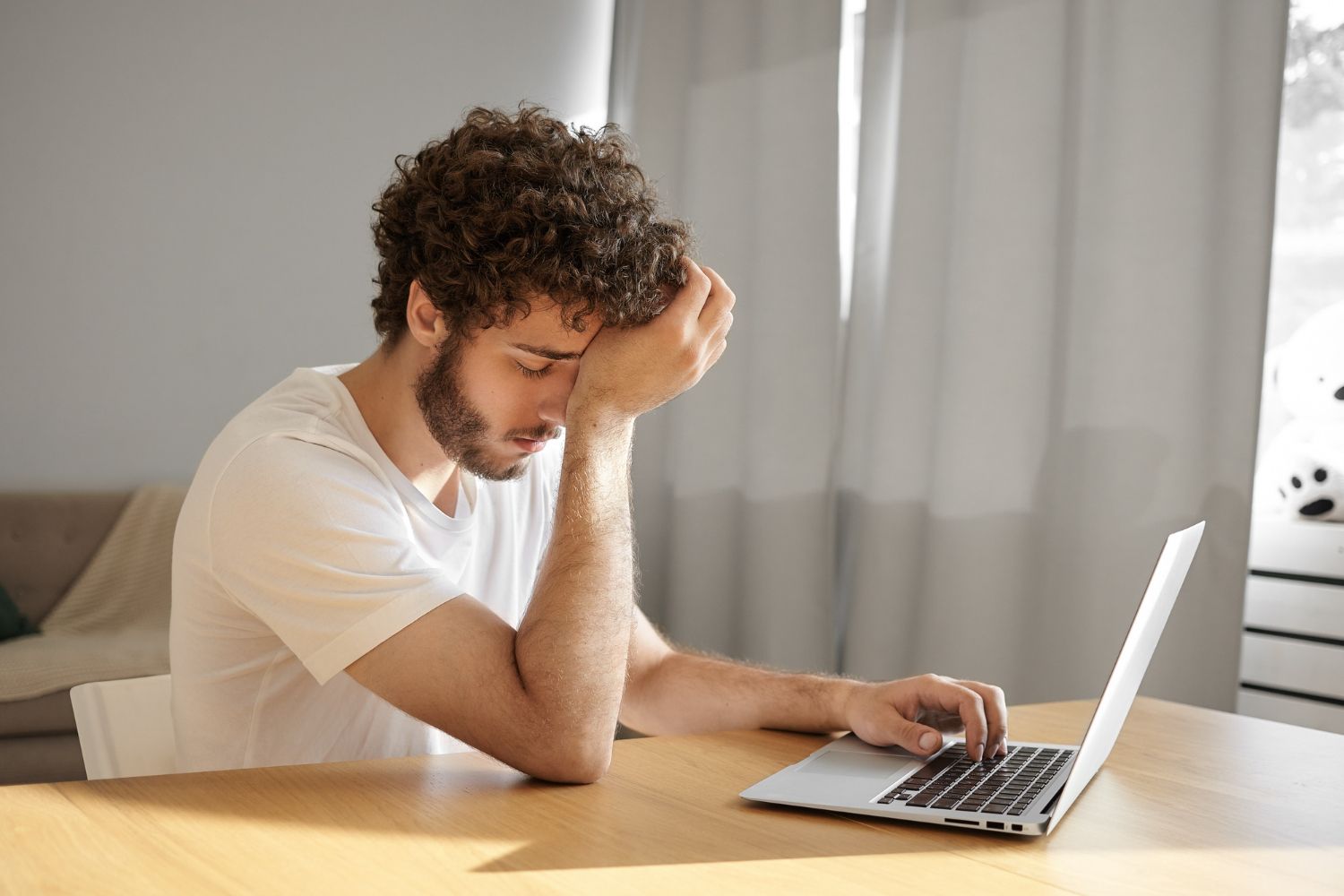 Man with curly hair, leaning head on hand, looking down at a laptop on a table.