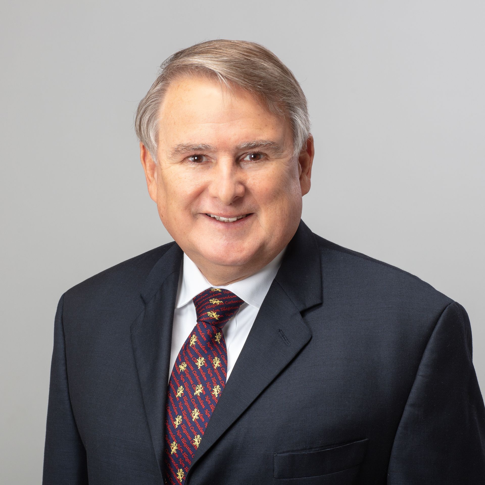 Man in suit smiling, looking at the viewer. Dark blue suit, patterned tie, gray background.