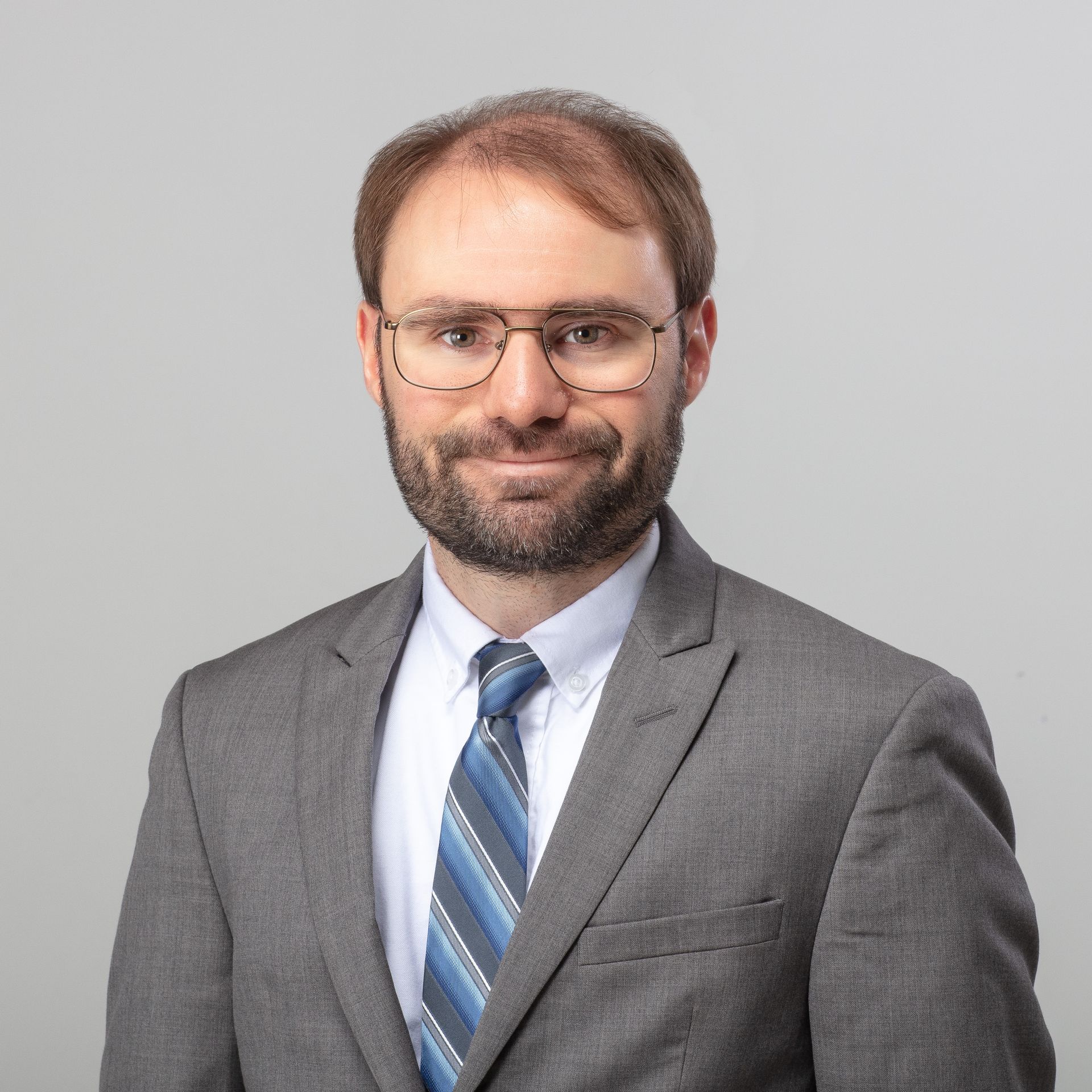 Man with beard and glasses wearing a gray suit and patterned blue tie, smiling in front of a gray background.
