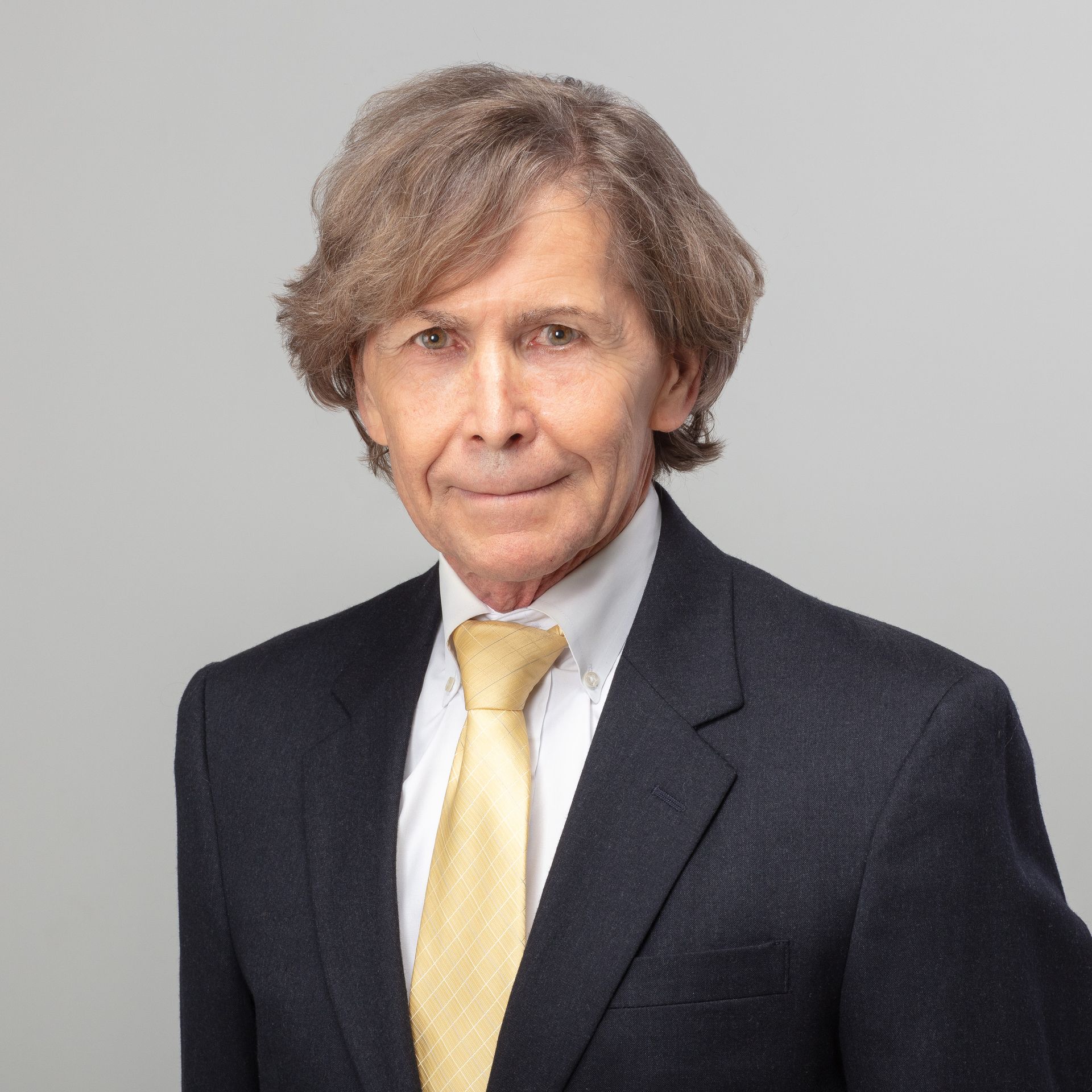 Man in suit with yellow tie smiles at the camera, neutral background.