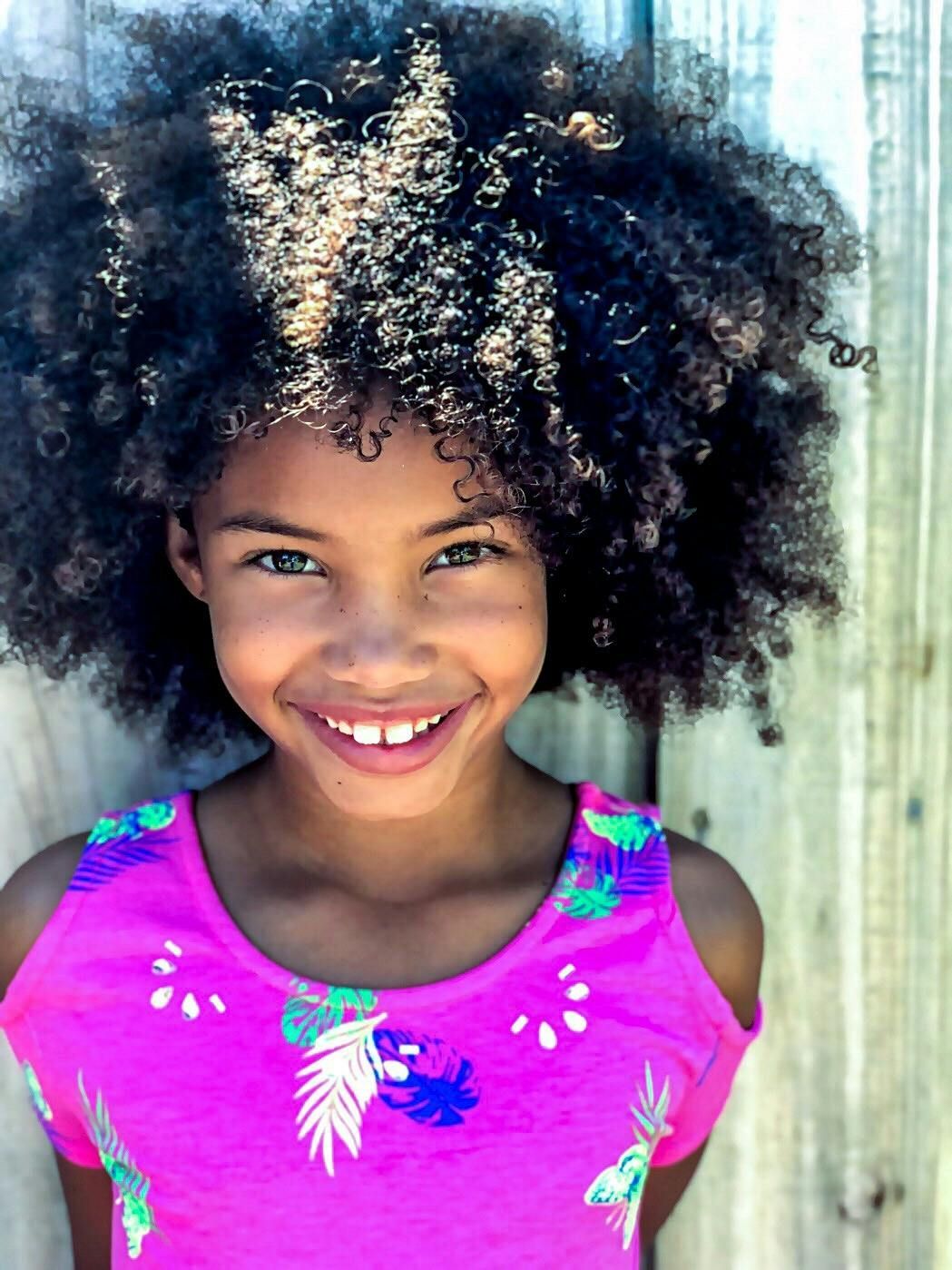 Girl with large, curly hair, smiles brightly; pink top, wooden fence backdrop.