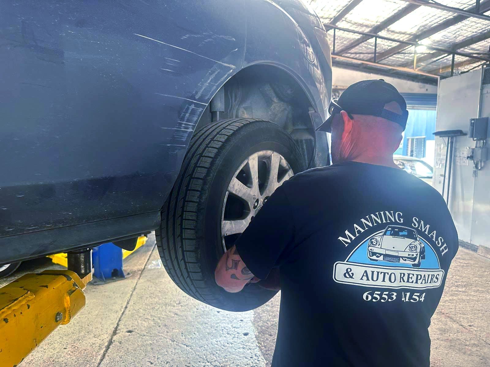 A man is working on the underside of a car in a garage. -  Manning Smash & Auto Repairs In Wingham, NSW