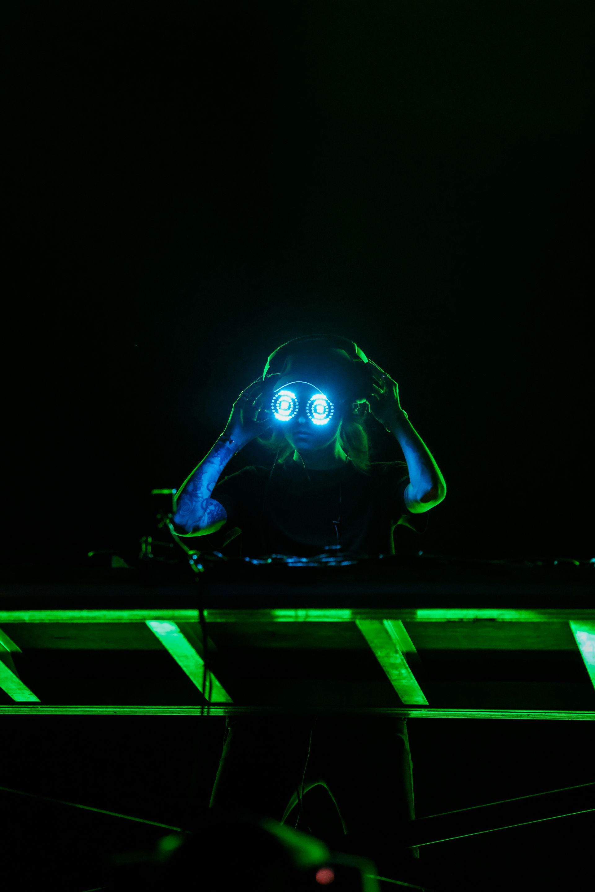 A dj is playing music in a dark room with green lights behind him.