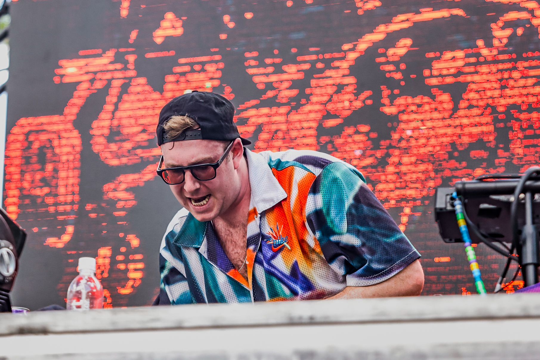 A man wearing sunglasses and a hat is standing in front of a large screen.