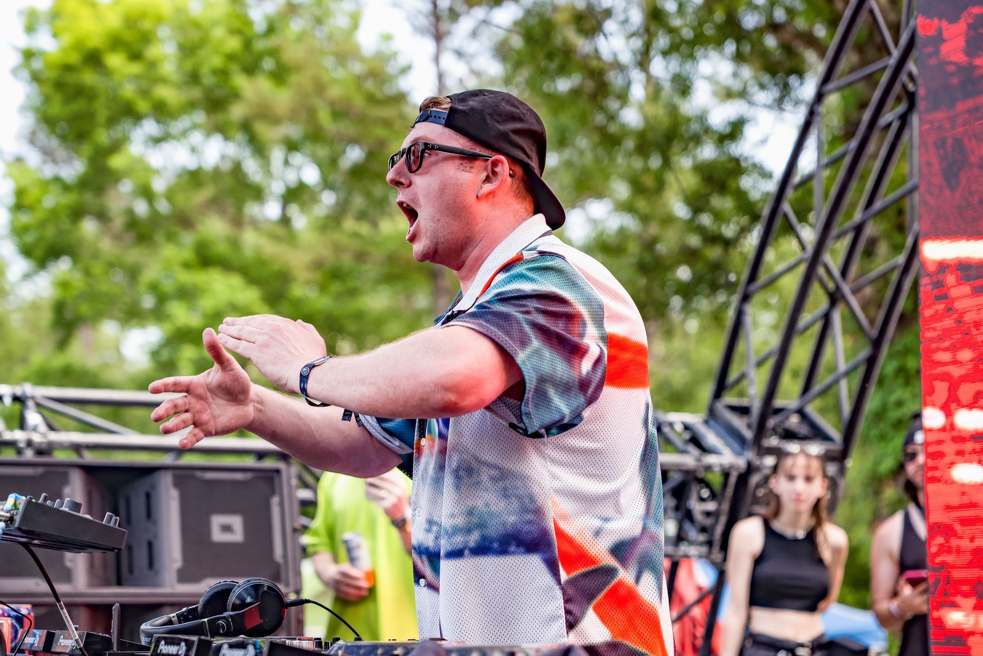 A man in a hat and sunglasses is standing in front of a dj booth.