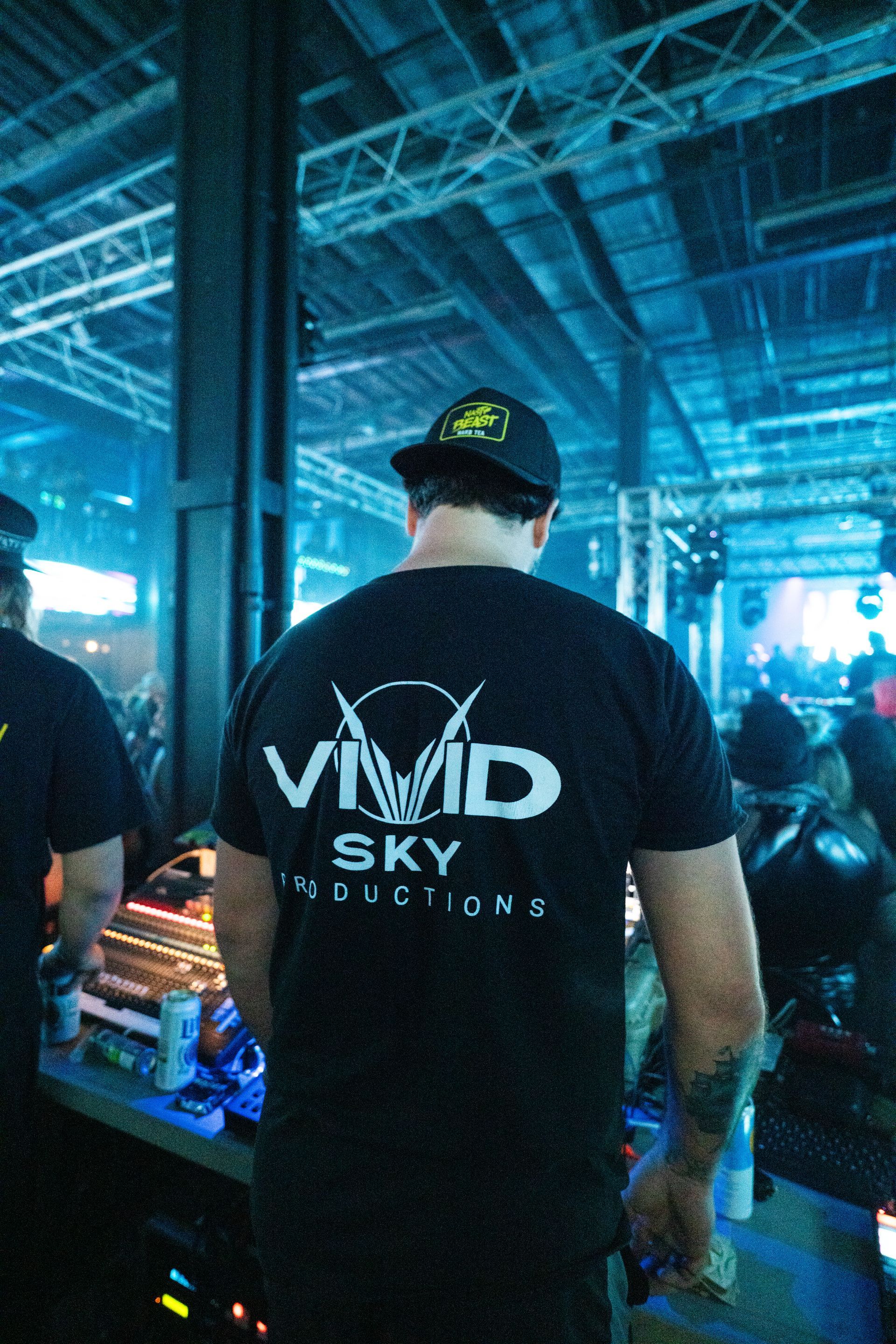 A man wearing a vivid sky t-shirt is standing in a dark room.