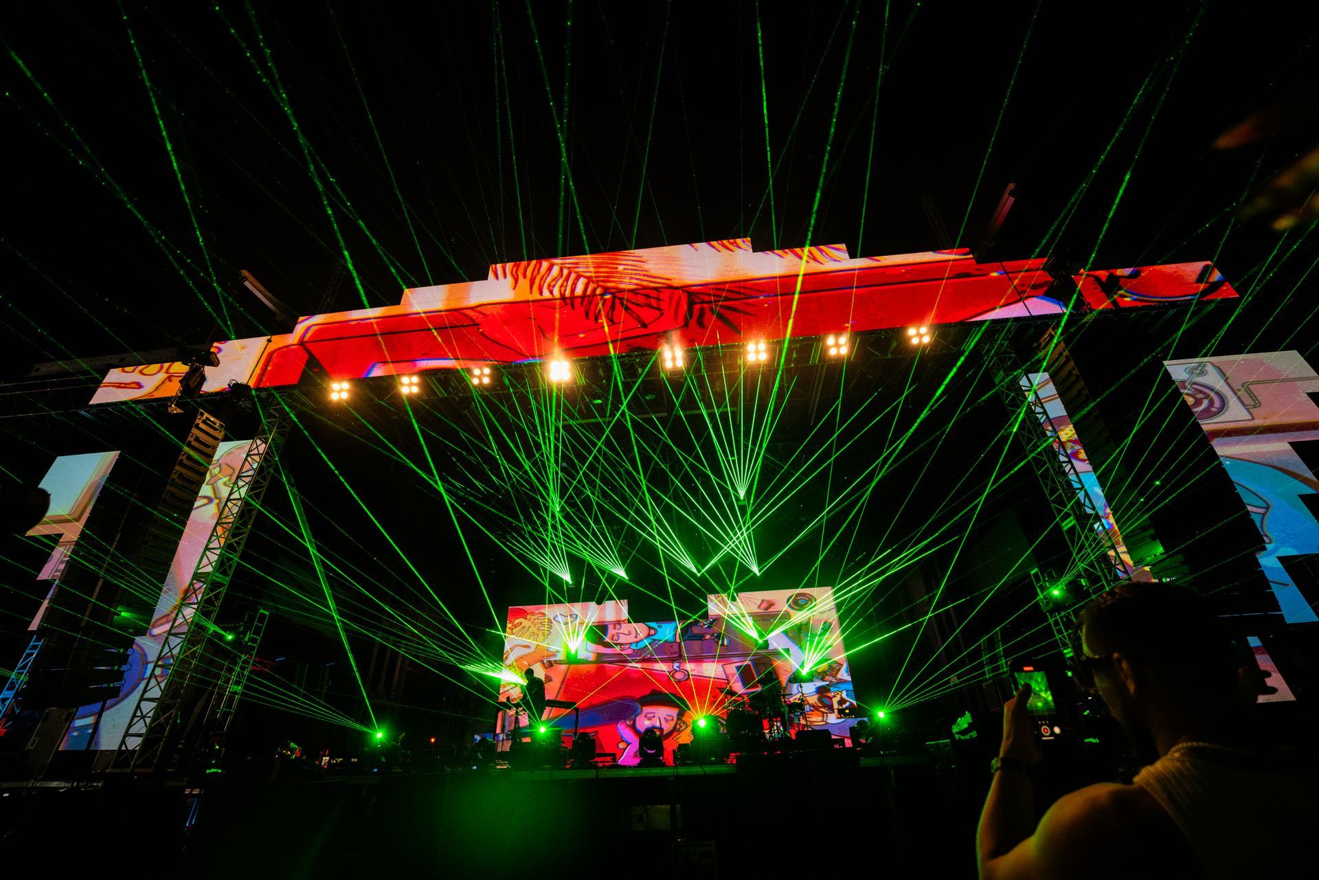 A man is taking a picture of a stage at a concert with green lights coming out of it.