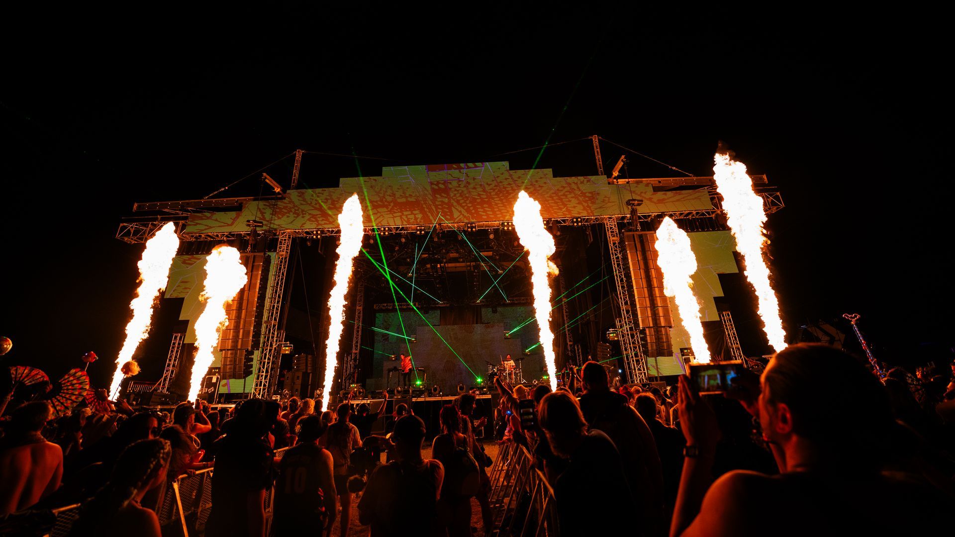 A crowd of people are watching a concert with fire coming out of the stage.