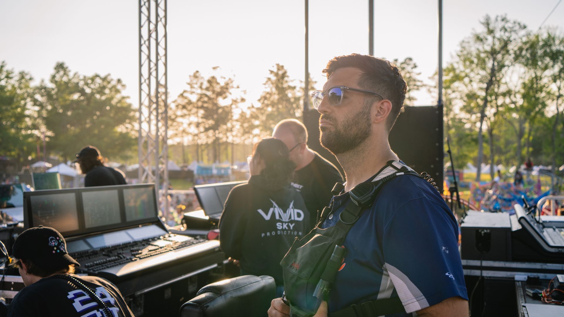 A man wearing sunglasses and a backpack is standing in front of a stage.