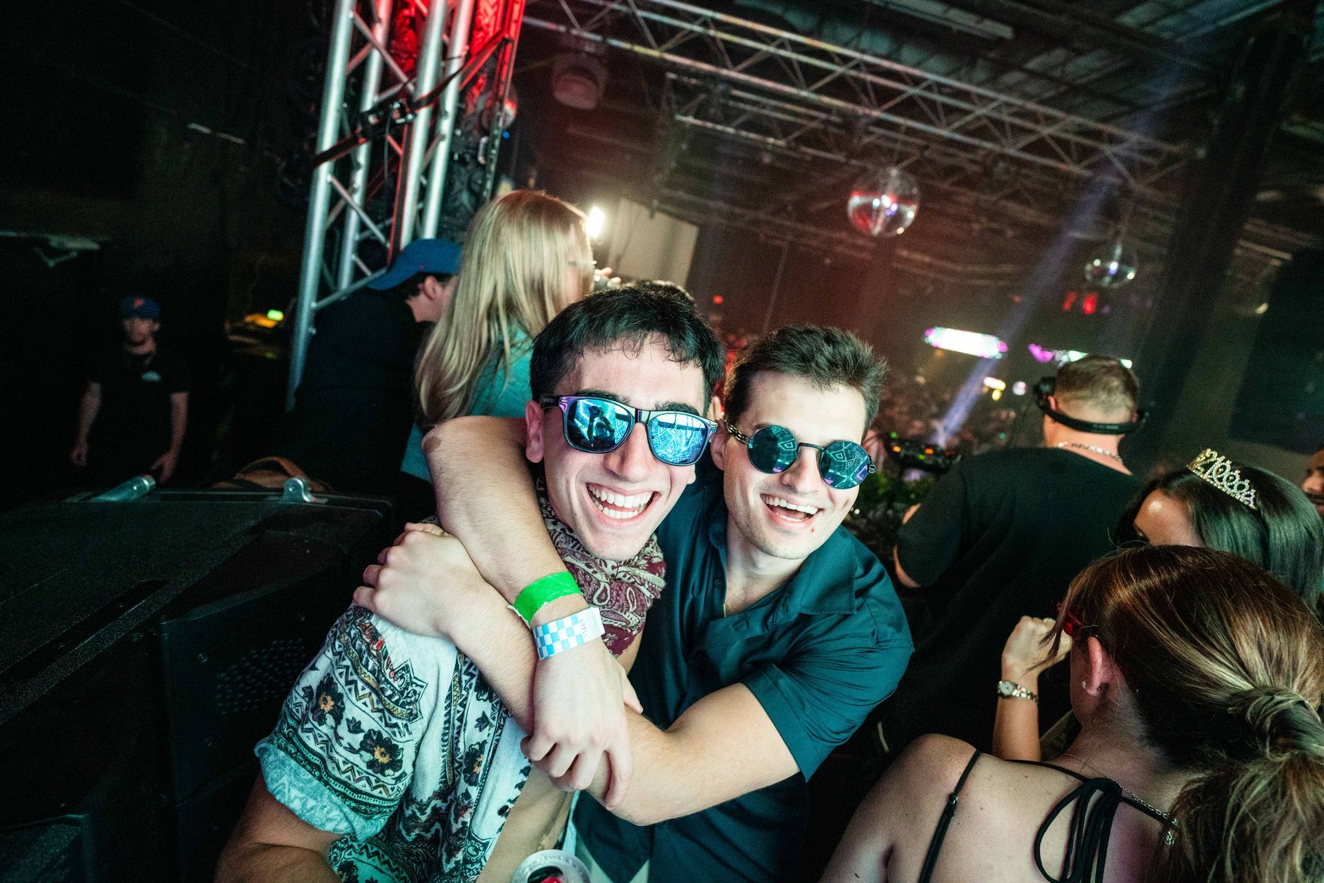Two men wearing sunglasses are hugging each other at a party.