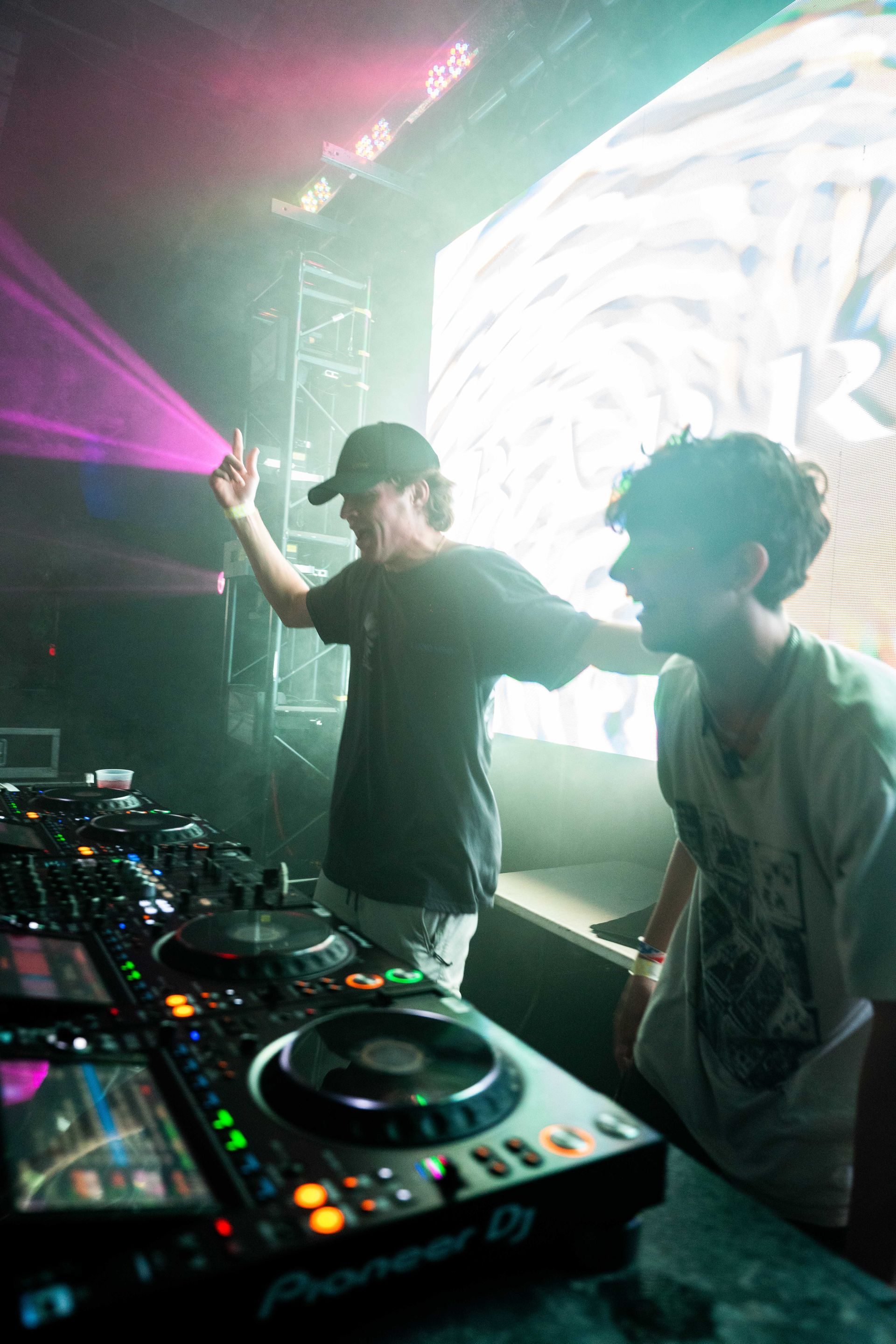 Two men are standing in front of a dj controller in a dark room.